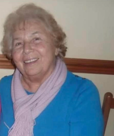 Smiling elderly woman with curly hair, wearing a blue top and purple scarf.