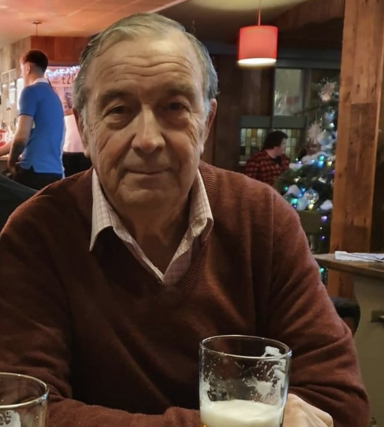 Elderly man in a brown sweater sitting at a table with a drink, smiling.