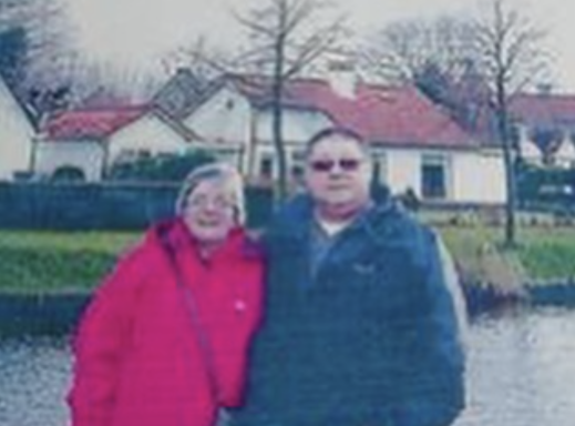 A couple by a river, wearing warm clothing, with houses and trees in the background.