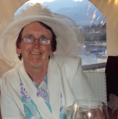 Smiling woman in a white hat and blazer, seated with a glass in a scenic setting.