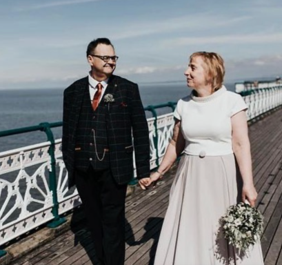 Couple walking hand in hand on a pier, dressed for a wedding, smiling at each other.