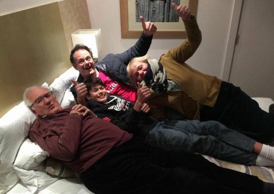 Four people playfully lying on a bed, laughing and making fun gestures.