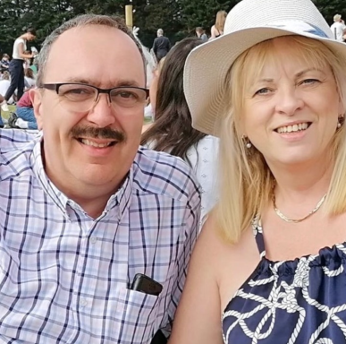 Smiling couple in casual attire, seated outdoors with people in the background.