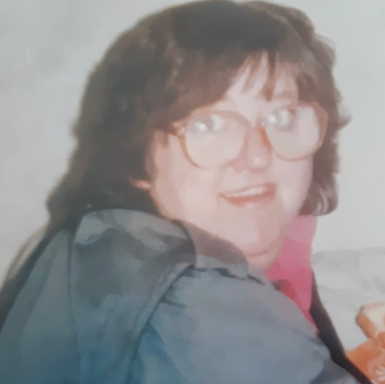 A woman with glasses and dark hair smiles, wearing a grey jacket and pink shirt.