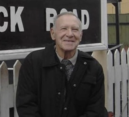 Smiling elderly man sitting beside a signpost for "Black Road".