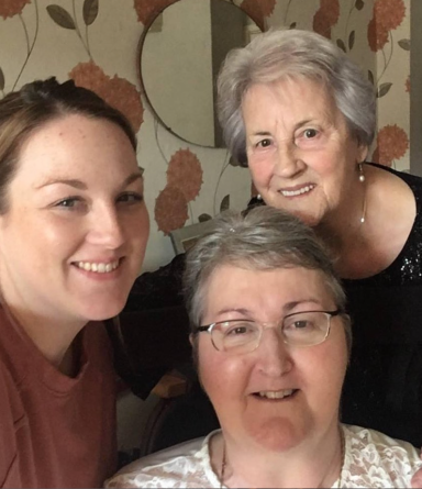 Three women smiling together, seated in a living room with floral wallpaper.