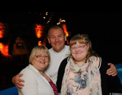 Three people smiling together, with a dark background and warm lights in the distance.