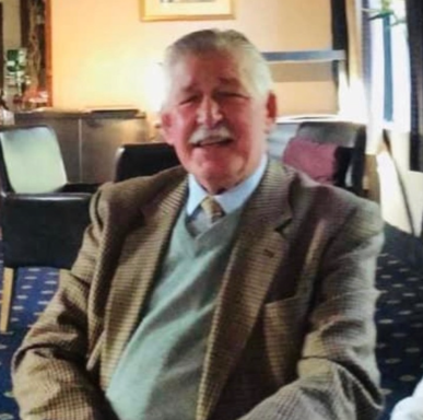 Elderly man sitting and smiling, wearing a suit jacket and a light blue sweater.