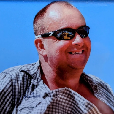Smiling man wearing sunglasses and a checked shirt against a blue sky.