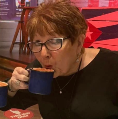 Older woman with short brown hair sipping from a blue cup in a lively café setting.