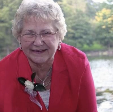 Smiling elderly woman in a pink cardigan, with a flower accessory, by a serene lake.