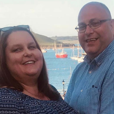 A smiling couple poses by the seaside with boats in the background.