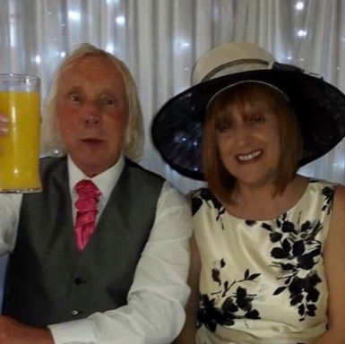 A man in a grey suit and a woman in a floral dress, both smiling, with drinks raised.