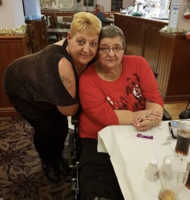 Two women pose together at a table in a restaurant, one seated in a wheelchair.