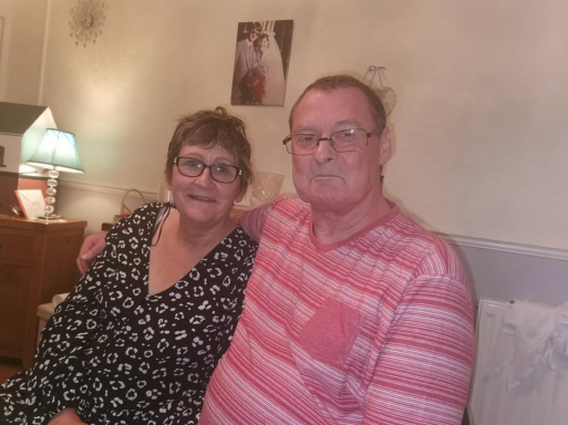 A middle-aged couple sitting together, smiling in a cozy indoor setting.