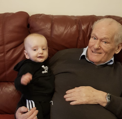 An elderly man sitting on a sofa with a smiling baby in a tracksuit.