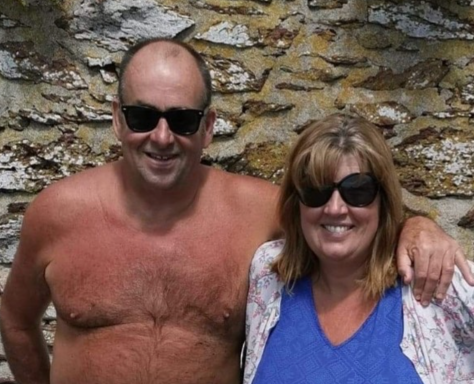 A man and woman smiling, wearing sunglasses, against a stone wall background.