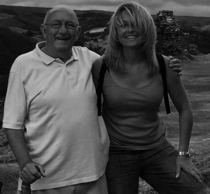 An older man and a woman stand together outdoors, smiling against a scenic mountainous background.