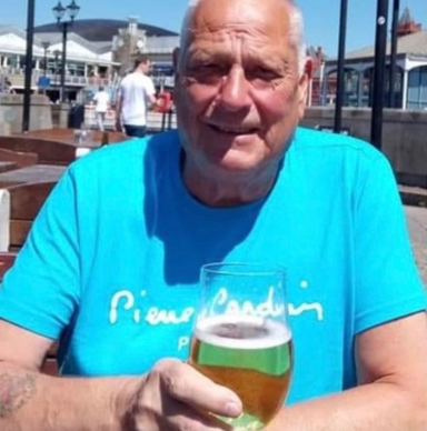 Older man in a blue shirt holding a glass of beer, seated outdoors.
