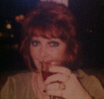 Woman with red hair holding a drink, smiling at the camera in a dimly lit setting.