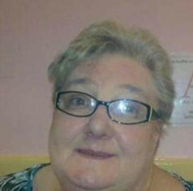 Elderly woman with glasses, smiling, against a pink wall background.