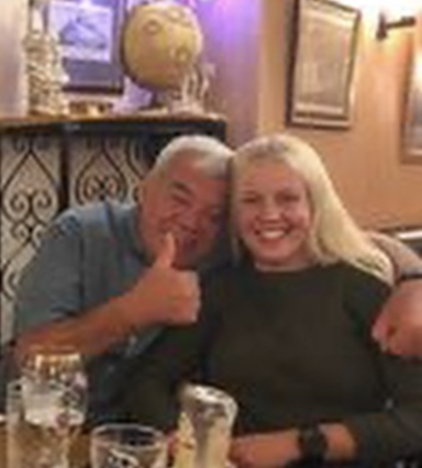 Two people smiling and posing together, one giving a thumbs up in a warm, cosy setting.