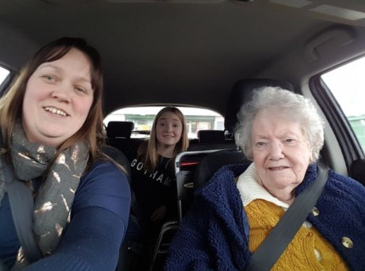 Three people seated in a car, smiling; a woman in a blue sweater, a girl, and an elderly woman.