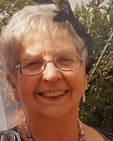 Smiling elderly woman with short grey hair, glasses, and a black decorative hairpiece.